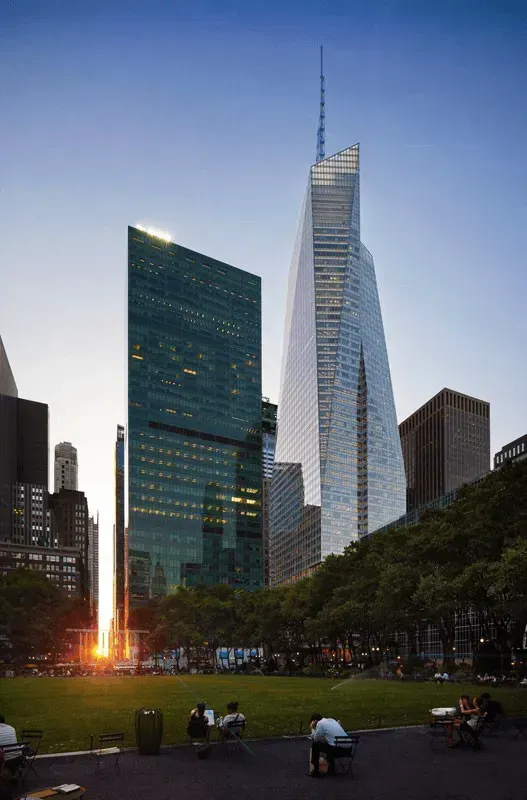 Evening city skyline with a tall glass skyscraper, a green park in front, and people sitting on the lawn.