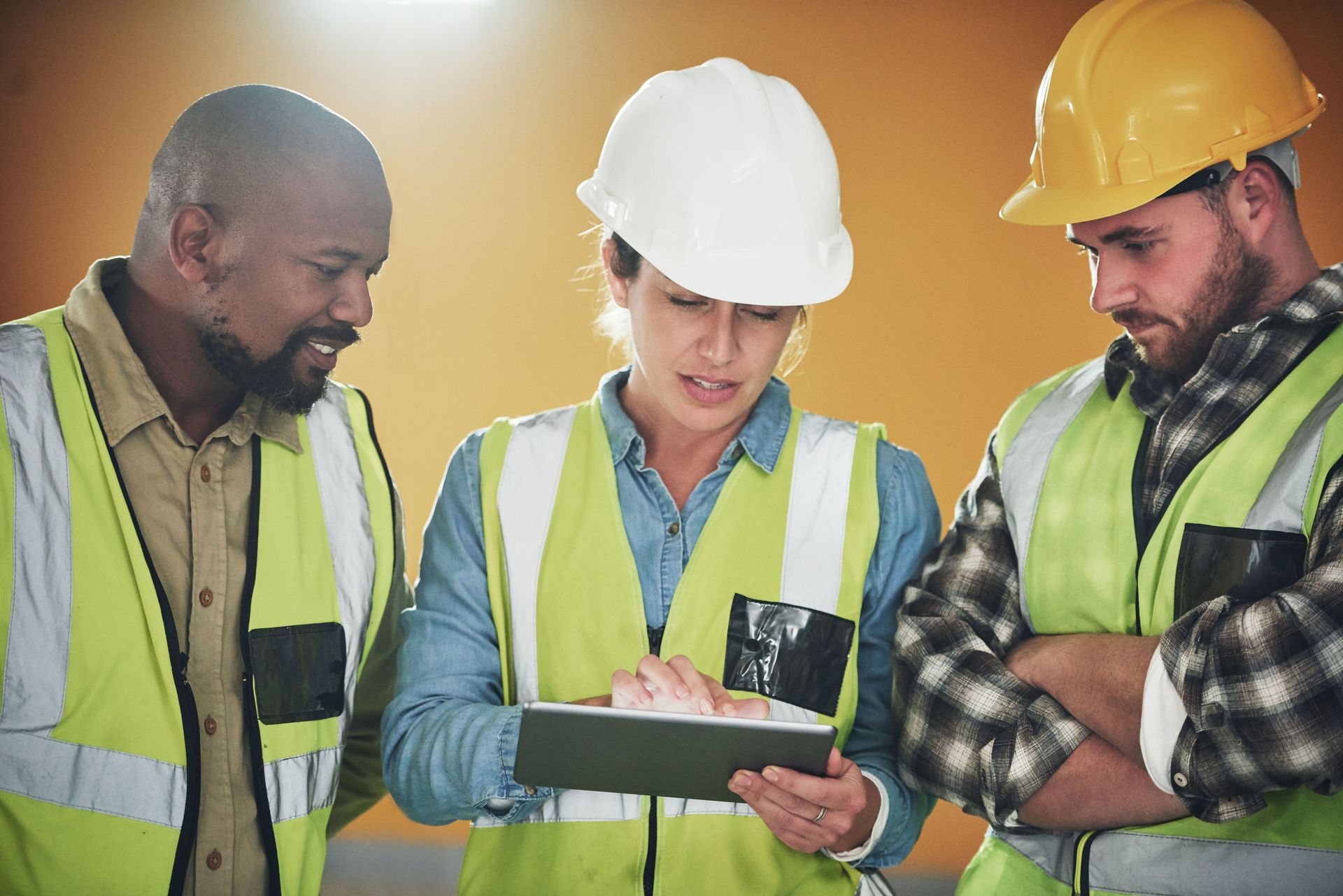 Three construction workers in hard hats and safety vests reviewing plans on a tablet at a work site