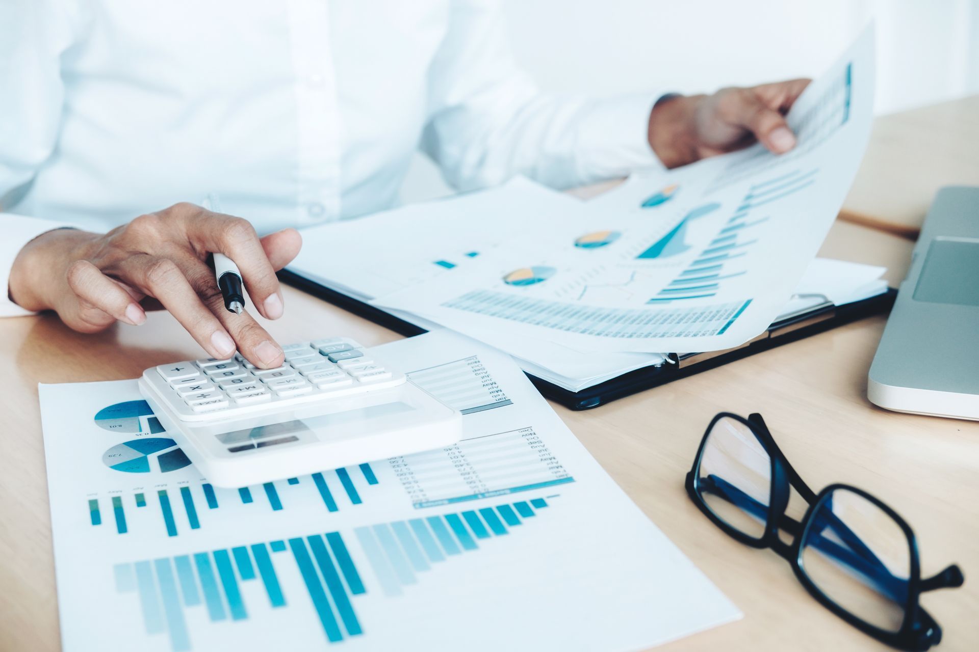 Person calculating financial data with a calculator, charts, and glasses.
