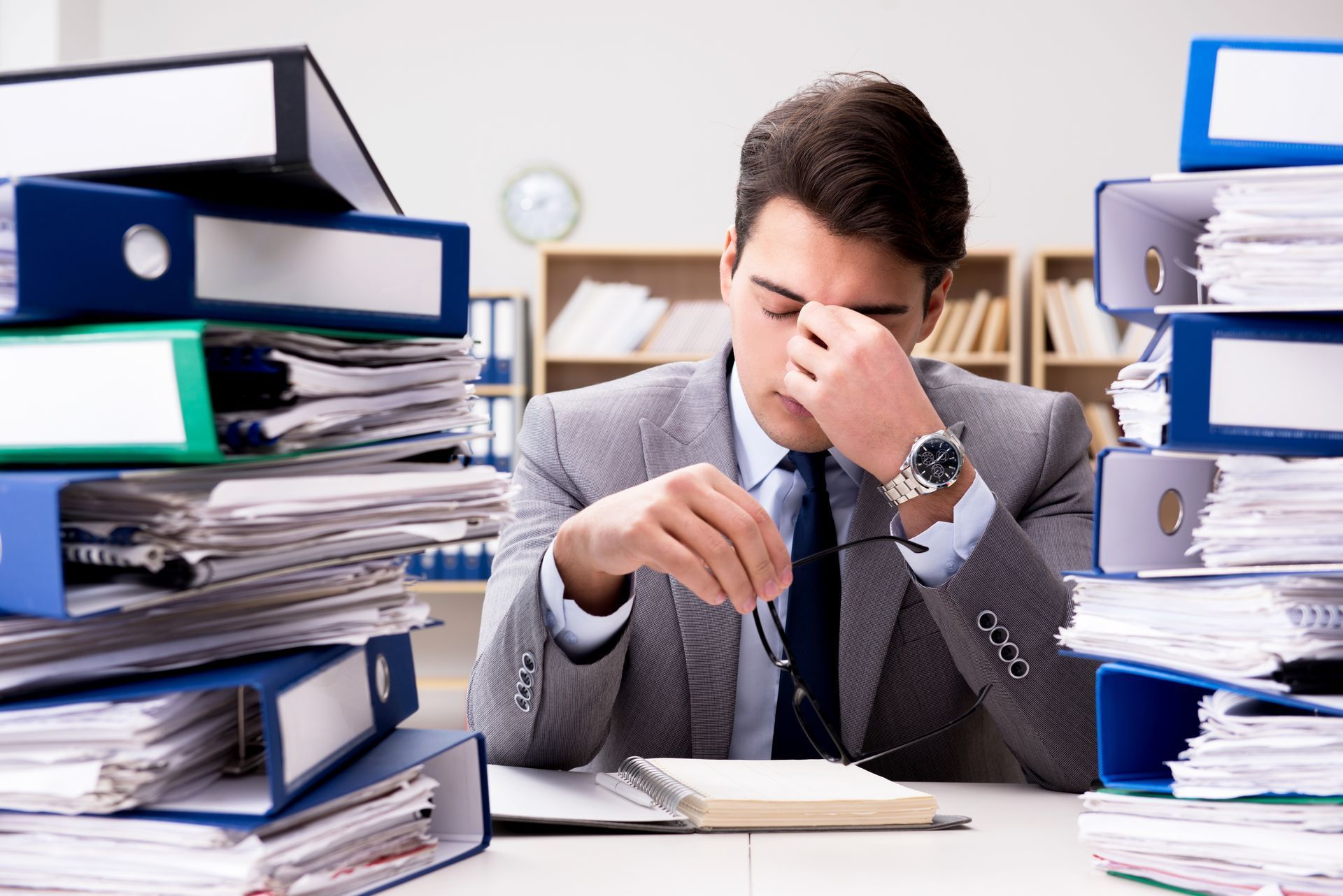Man in suit overwhelmed by paperwork, rubbing eyes in a cluttered office.