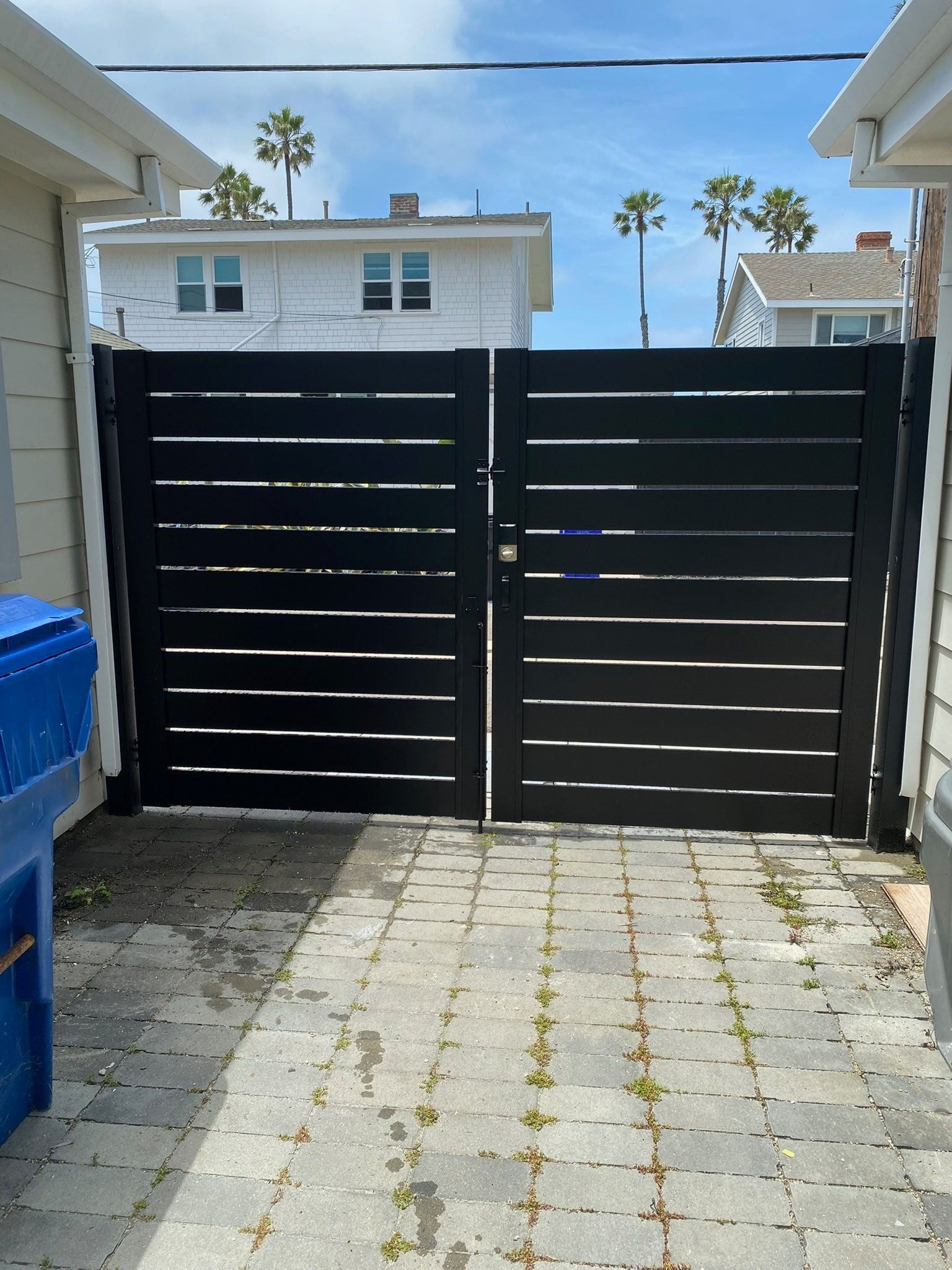 Black horizontal slat gate between two buildings, on a brick paved area, with blue trash bin.