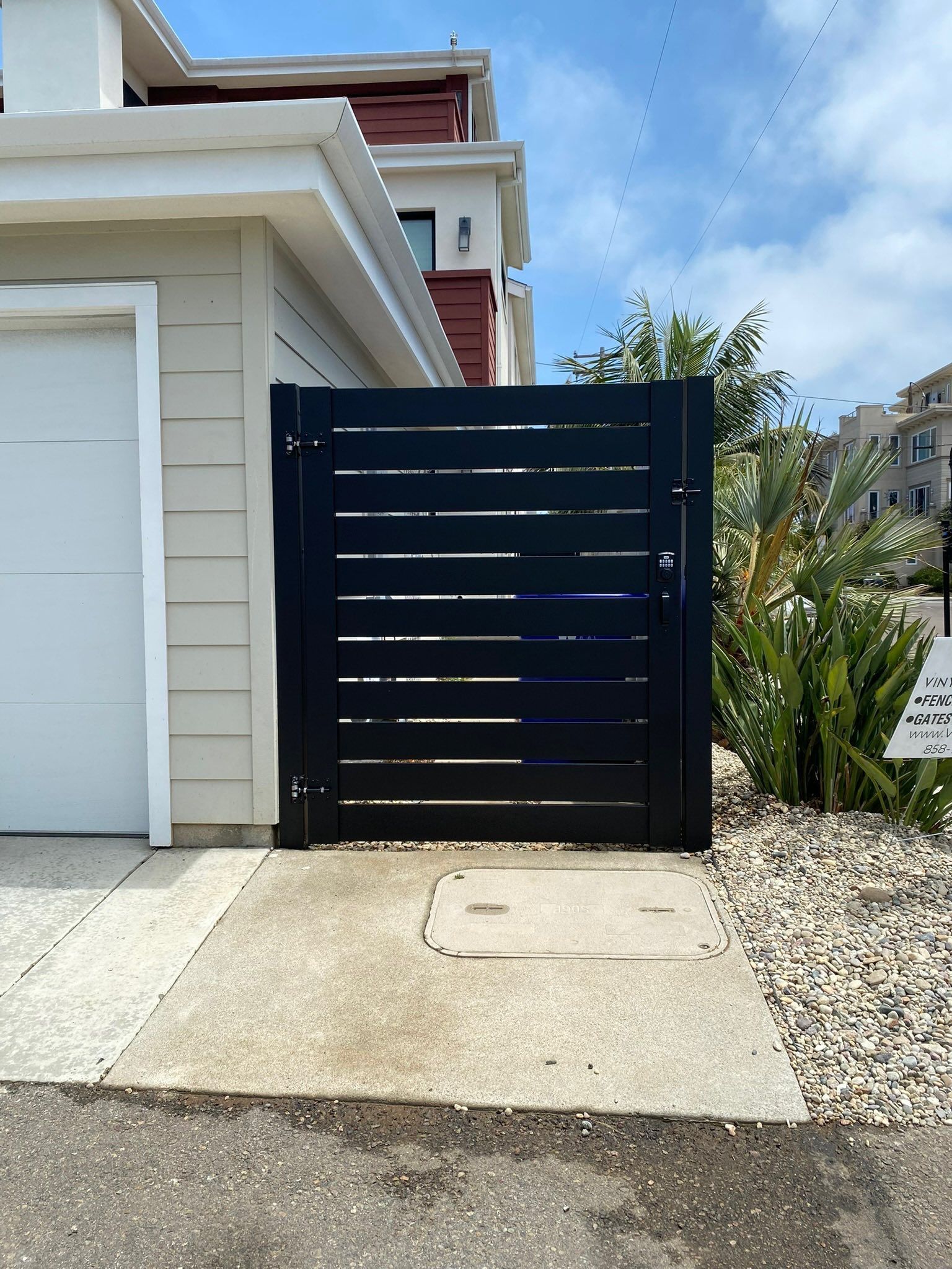 Black horizontal slat gate next to a garage and concrete path with a gravel bed and blue sky.