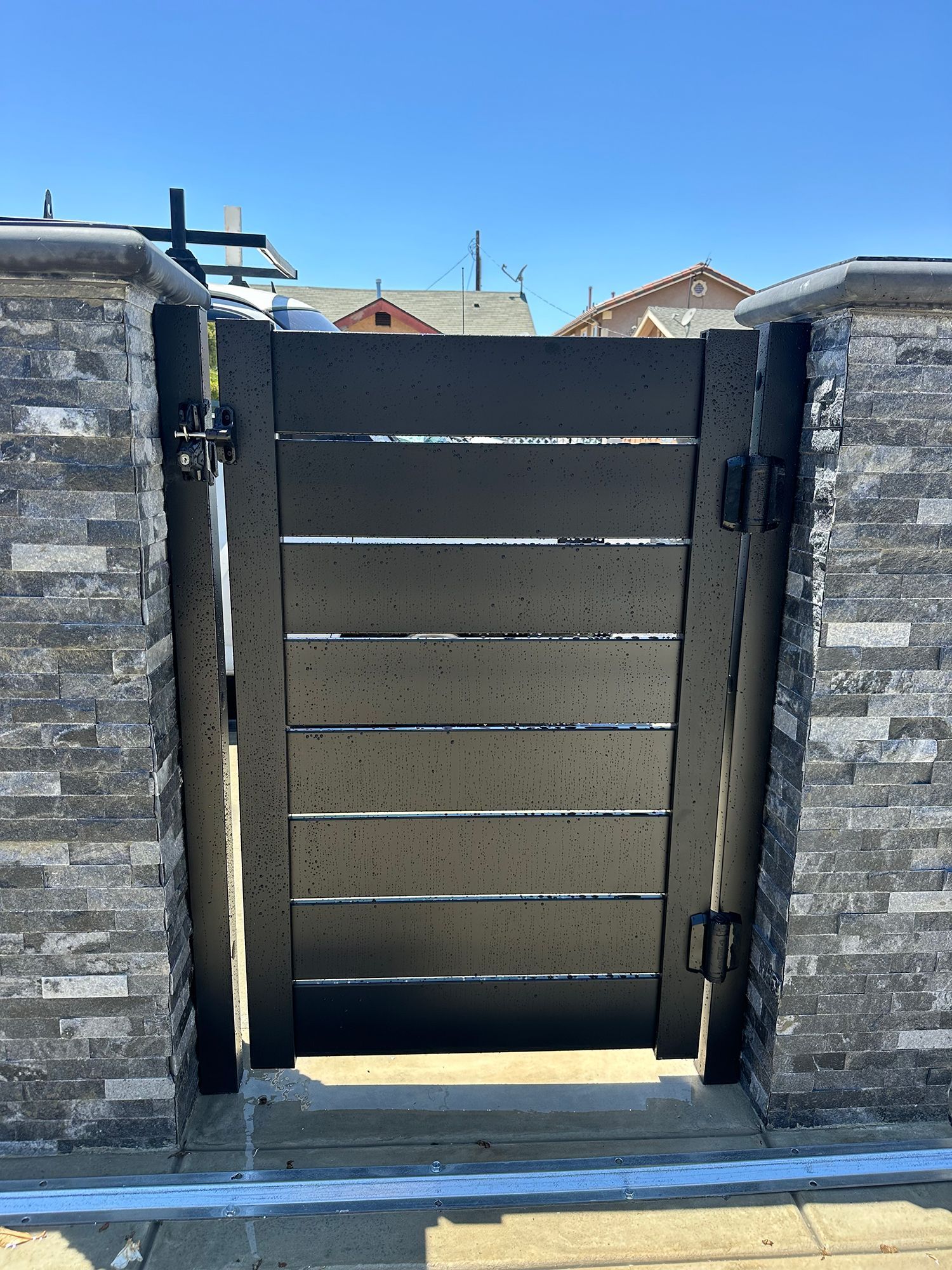 Black gate with horizontal slats, framed by stone columns, against a blue sky.