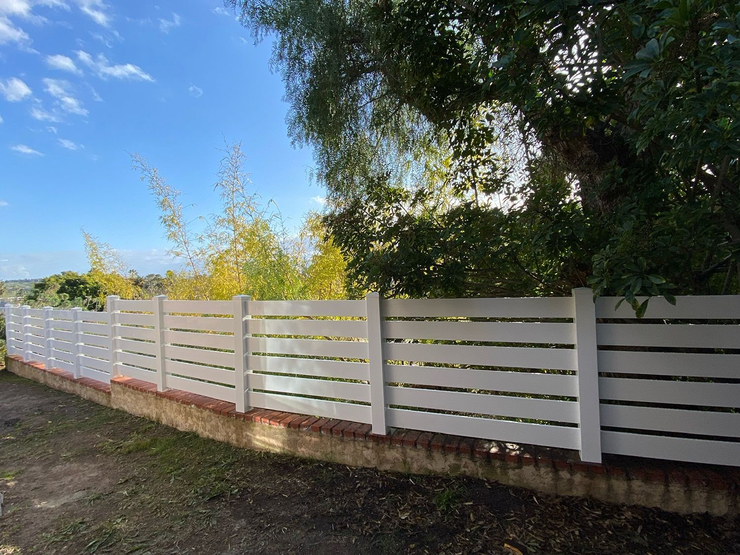 White horizontal slat fence on a low brick wall, against a backdrop of trees and a blue sky.