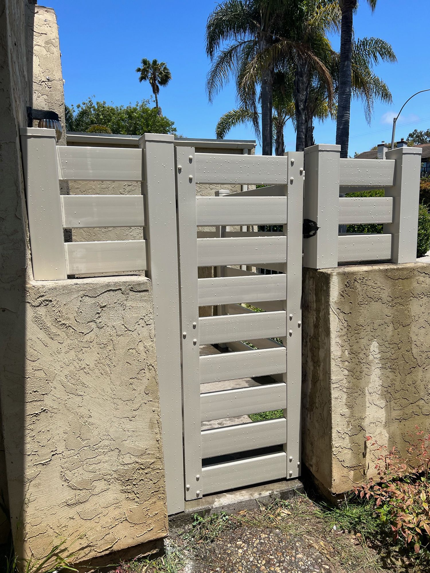 Gray slatted gate in a stucco enclosure, with palm trees in the background under a blue sky.