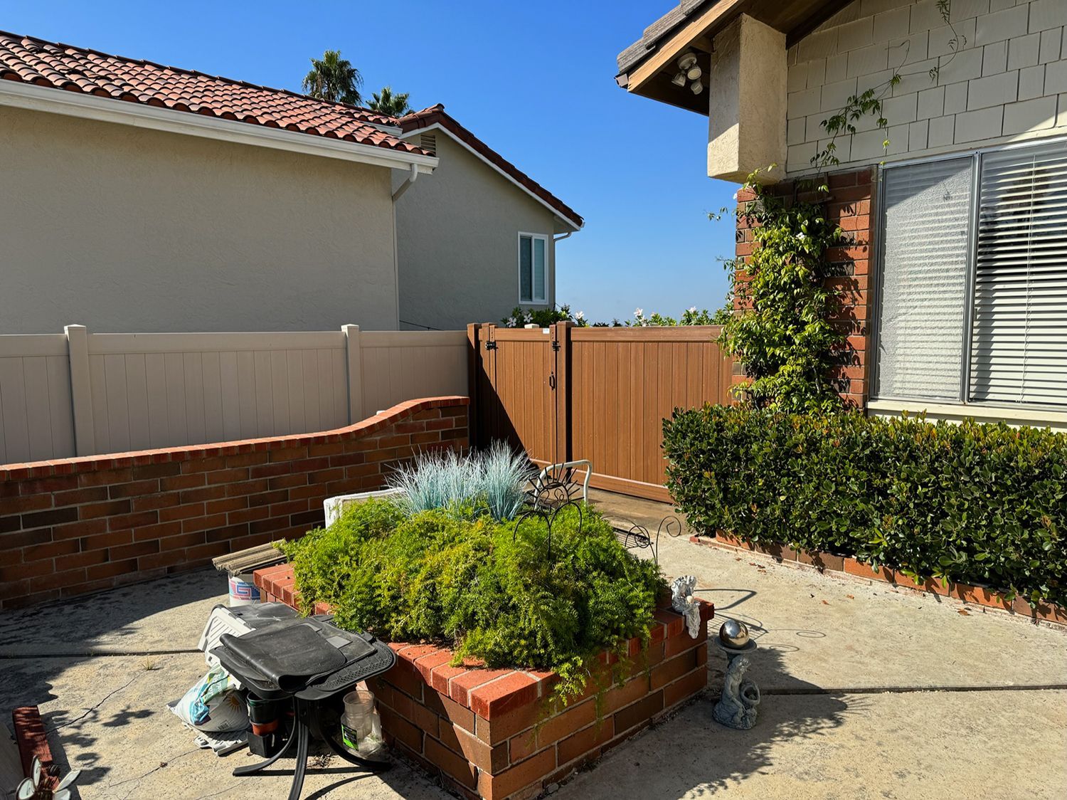 Backyard with brick planter, brown gate, beige fence and house, plants, and sunny sky.