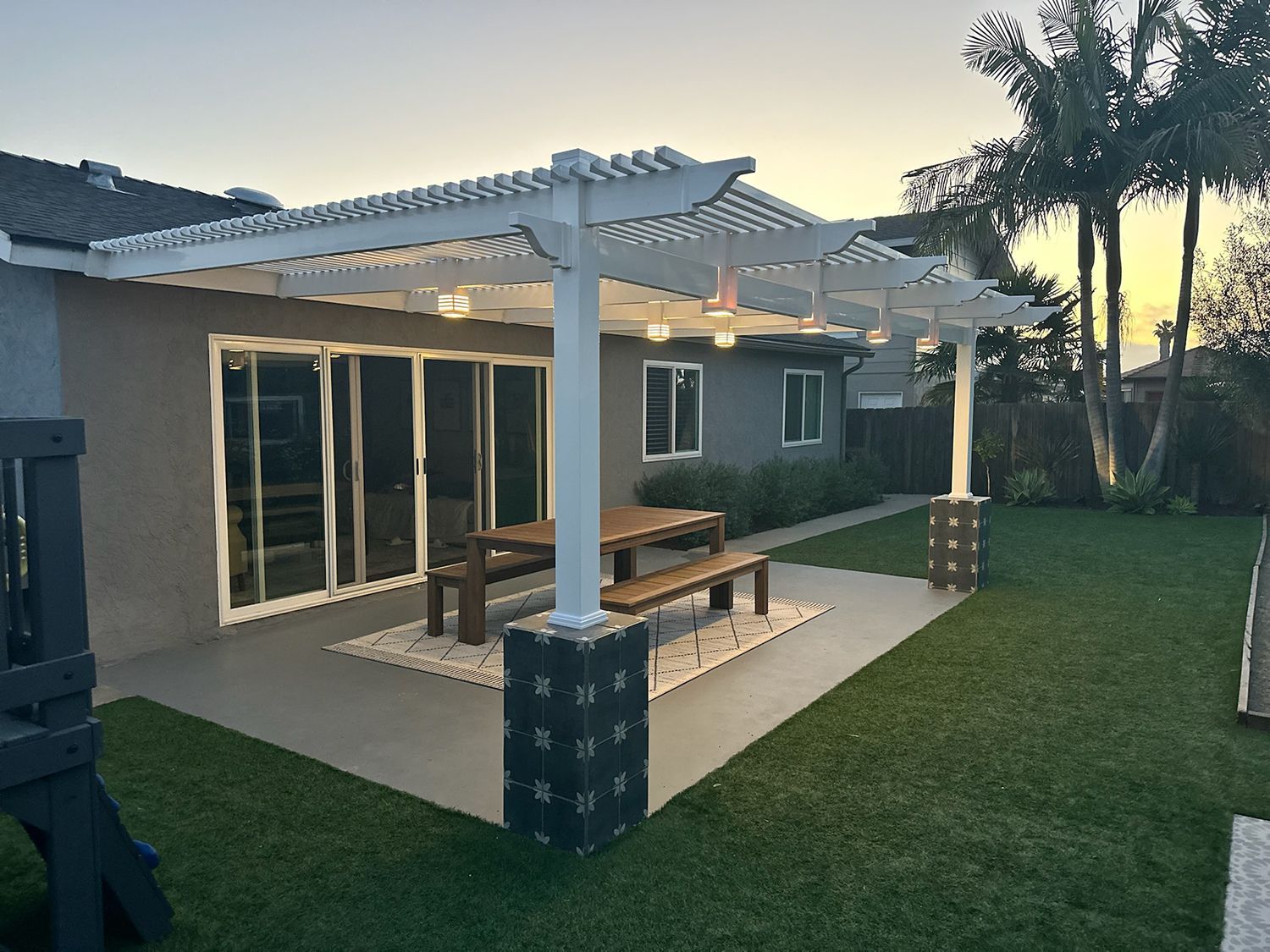 Patio with white pergola, table, benches, and string lights next to a house and green lawn.