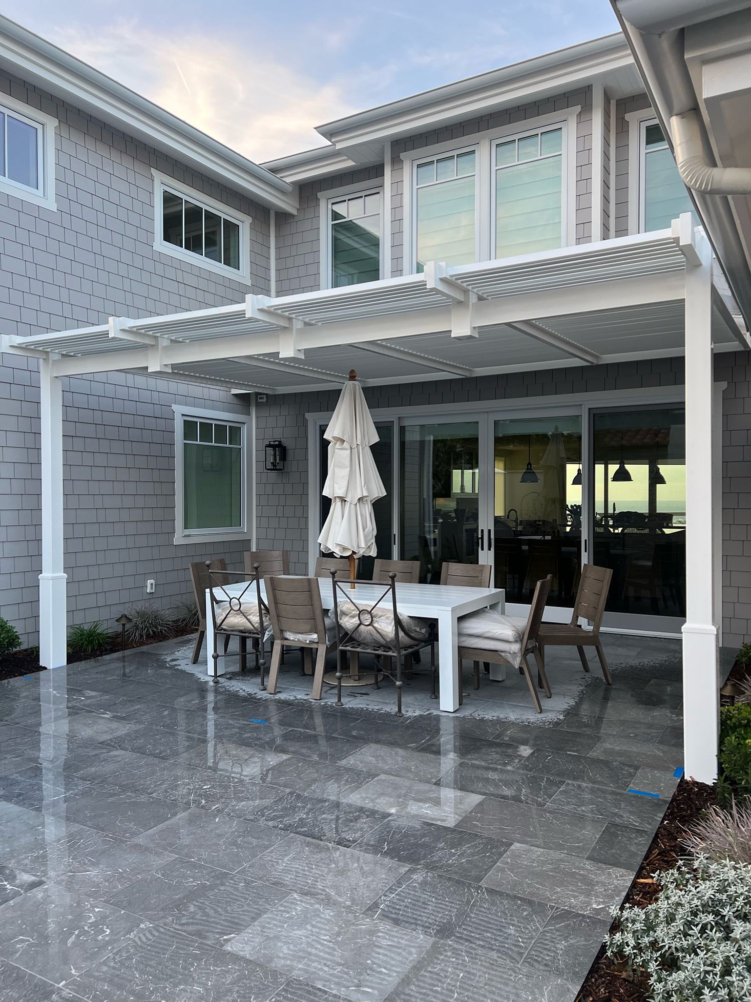 Gray patio with outdoor dining set, under a white pergola, next to a house.