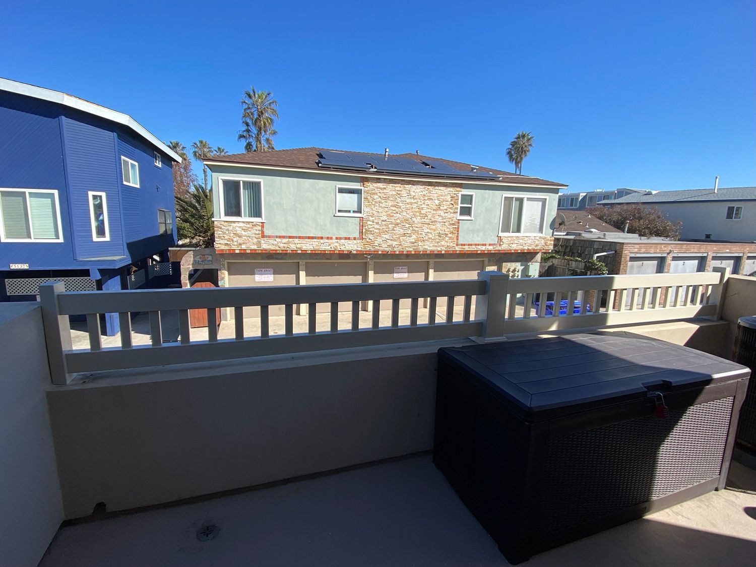 Patio with beige railing, wicker furniture, and a blue-painted building on a sunny day.