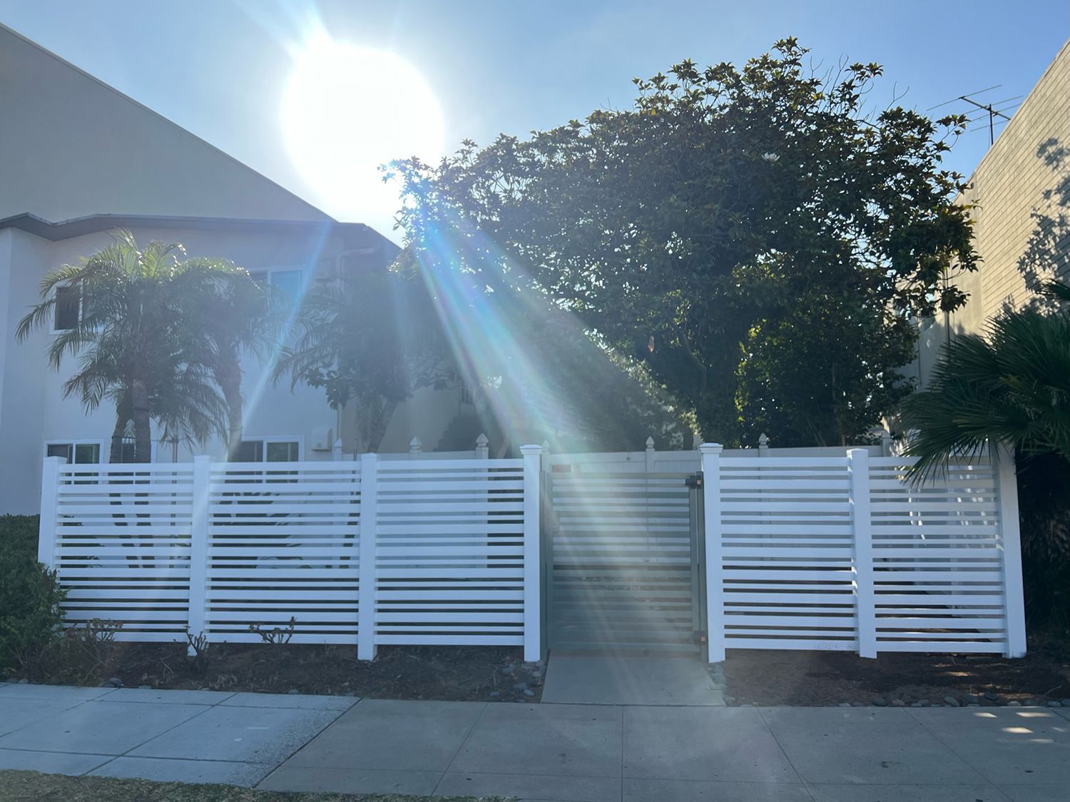 White horizontal slat fence with a gated entrance in front of a building, bright sunlight.