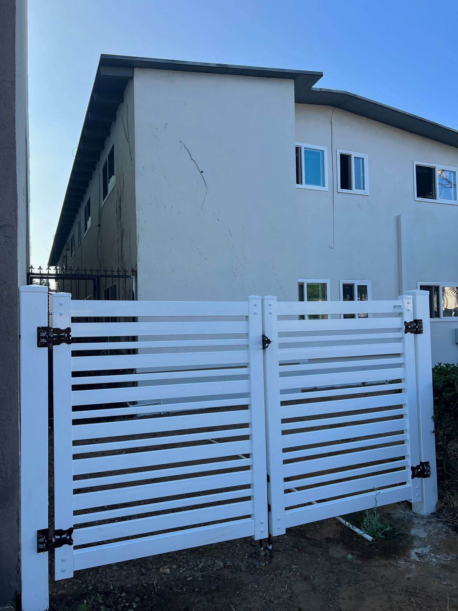 White slatted gate in front of a two-story stucco building with windows, under a clear sky.