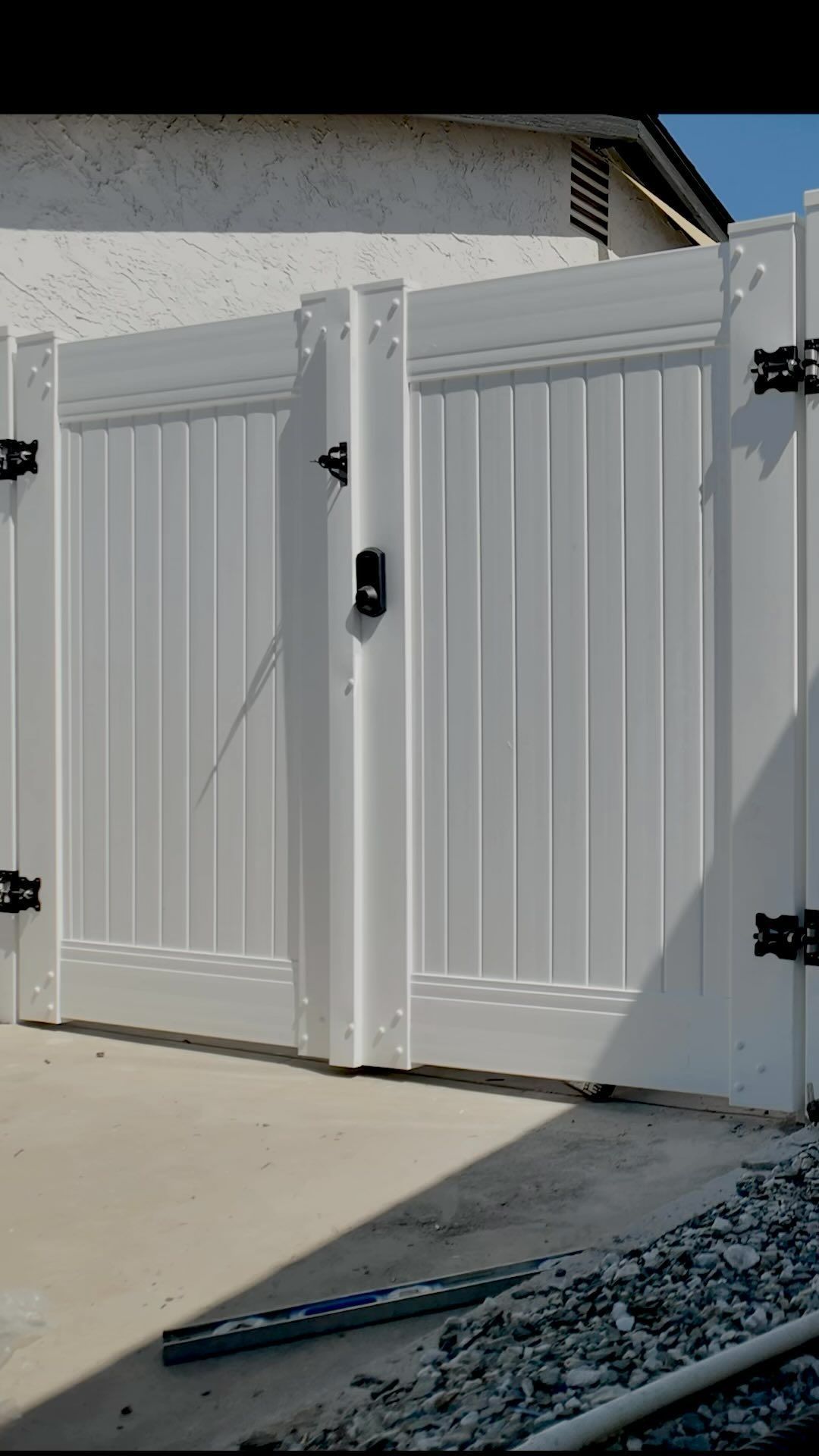 White double gate with black hinges and a lock, set on a concrete and gravel surface.