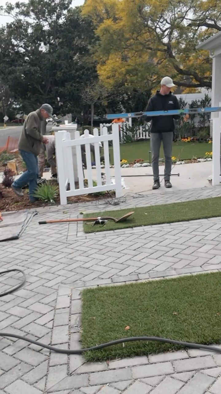 Two men installing a blue rail fence near a white picket fence and artificial turf on a brick patio.