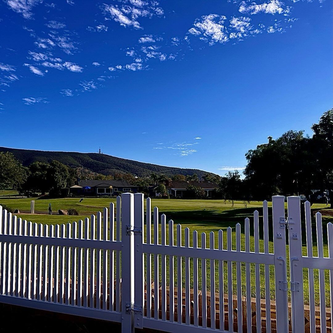 White picket fence frames a green field, with a mountain and blue sky dotted with clouds in the background.