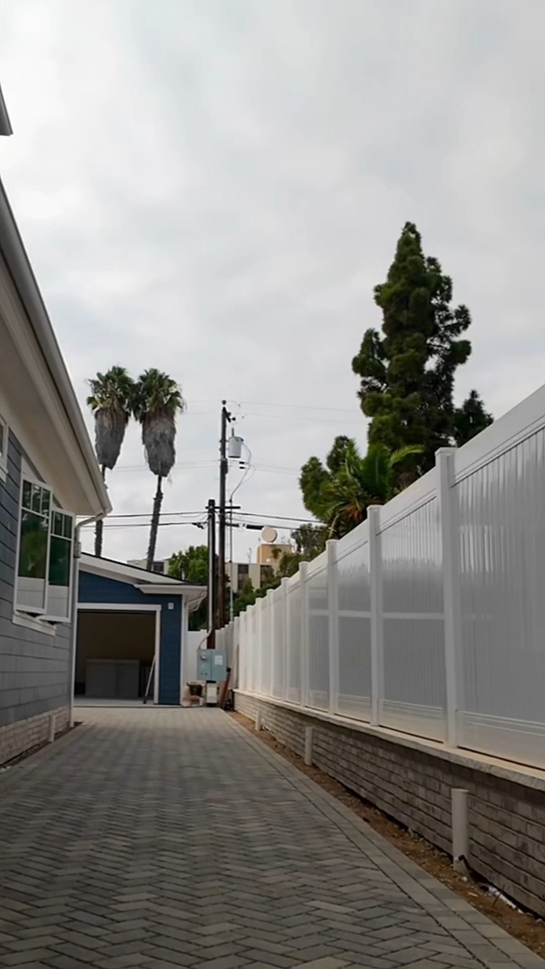 A brick-paved alleyway between a blue house and white fence under a cloudy sky, with palm trees in the distance.