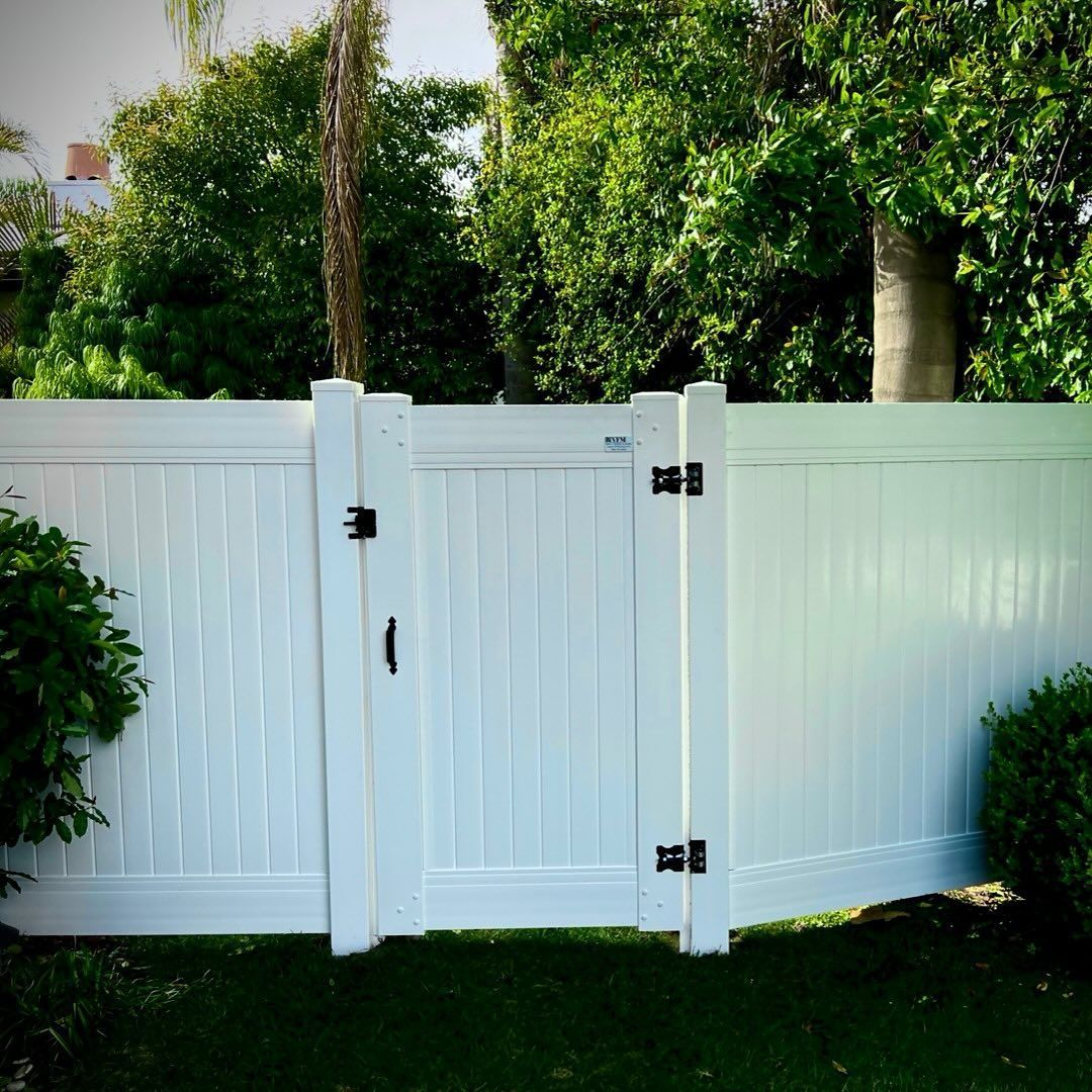 White vinyl fence with a gate, set in front of green trees and shrubs.