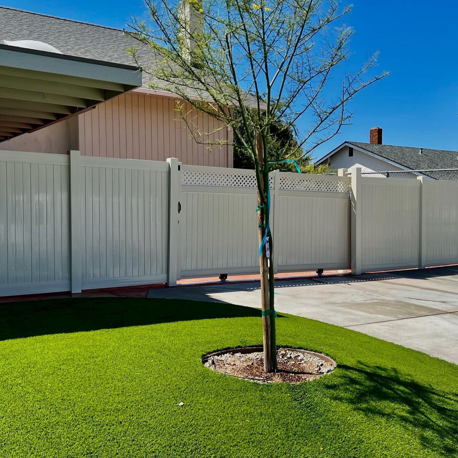 A cream-colored fence encloses a yard with green artificial turf. A young tree stands in the grass.