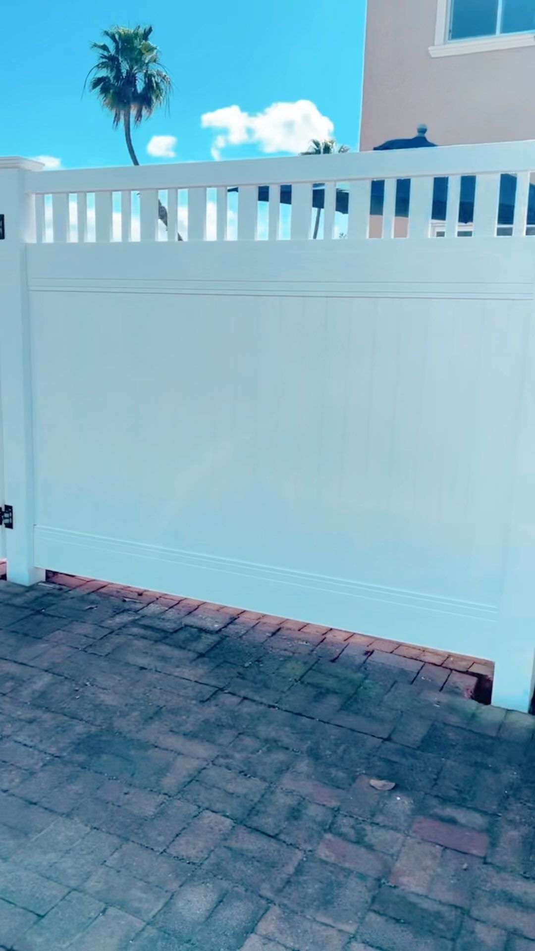 White fence with brick pathway, palm tree in background, blue sky.