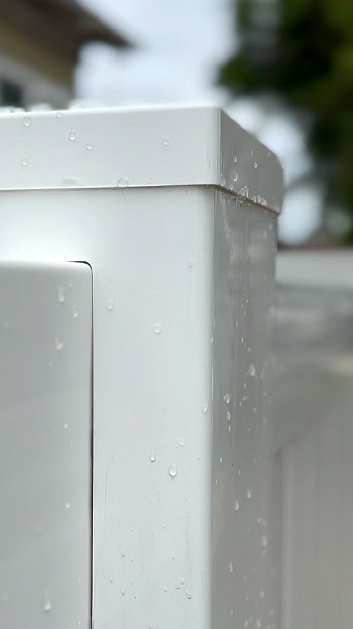 White fence post with water droplets, against a blurred background of a building and greenery.