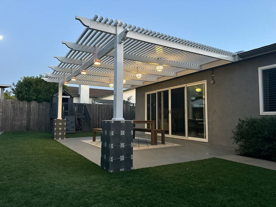 White pergola with string lights over a patio, gray house in background, green lawn.