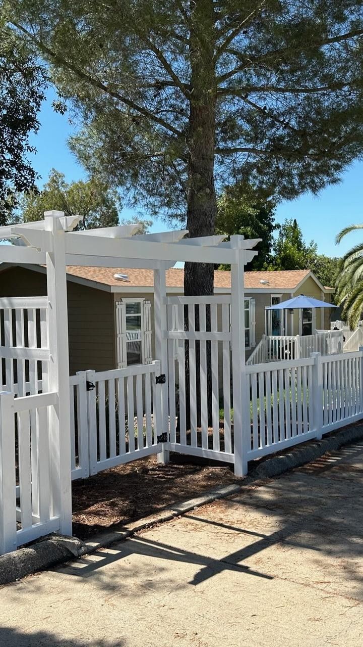 White picket fence surrounds a house; a pergola and trees are in the background on a sunny day.