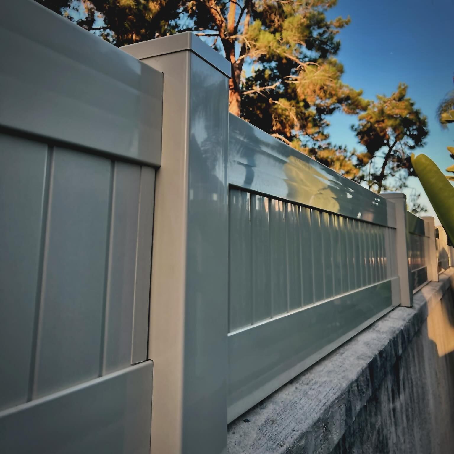 Gray vinyl fence atop a concrete wall, with a tree background.