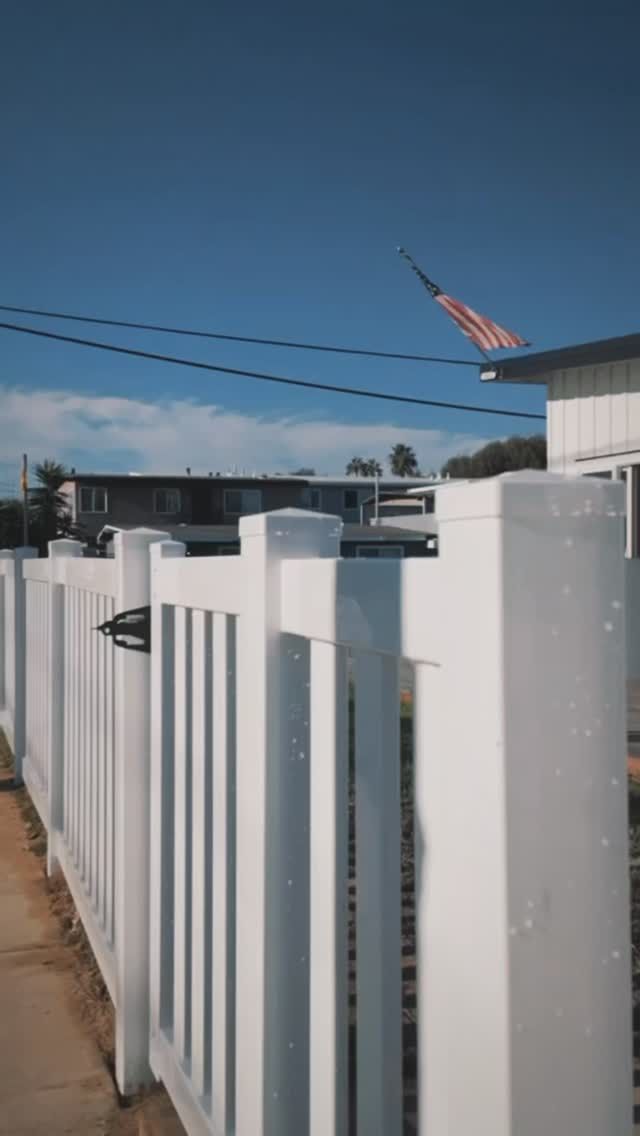 White picket fence with American flag waving in the background on a sunny day.