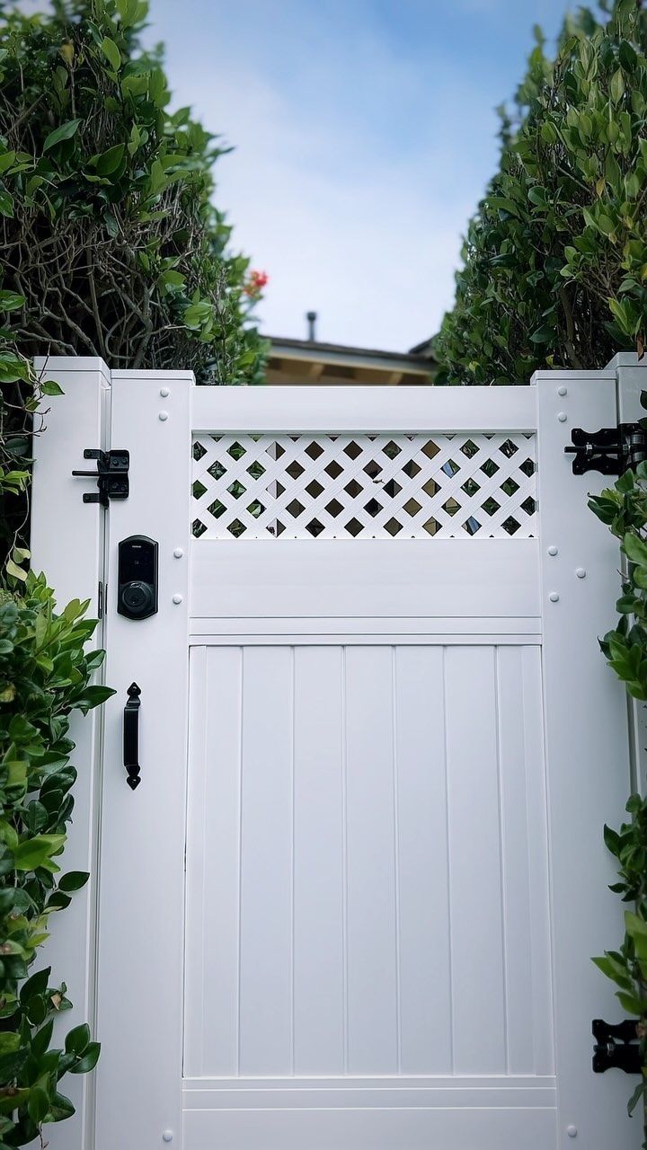 White gate with lattice detail, flanked by green hedges, leads to a house under a blue sky.