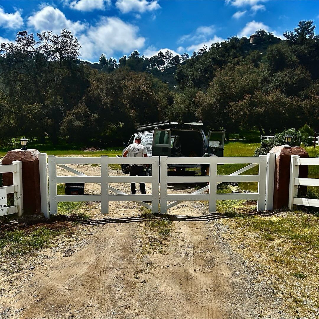 White gate and columns with a van, person, and trees in a field on a sunny day.