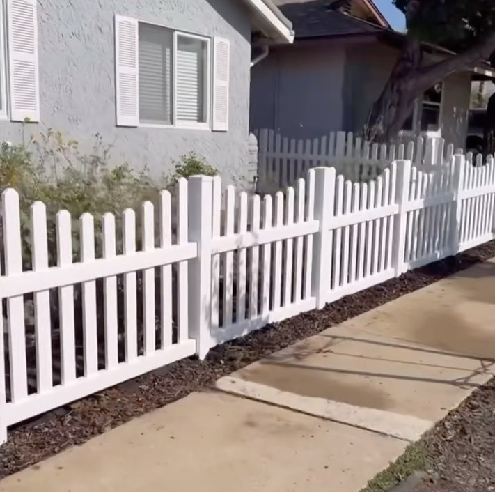 White picket fence in front of a light gray house with a sidewalk, tree, and window with shutters.