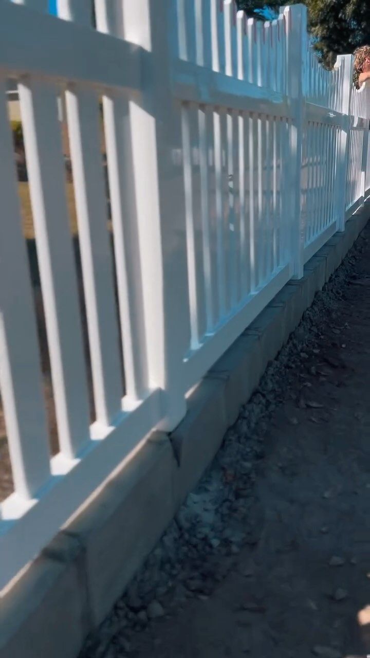 White picket fence along a concrete base, casting shadows on a dirt path.