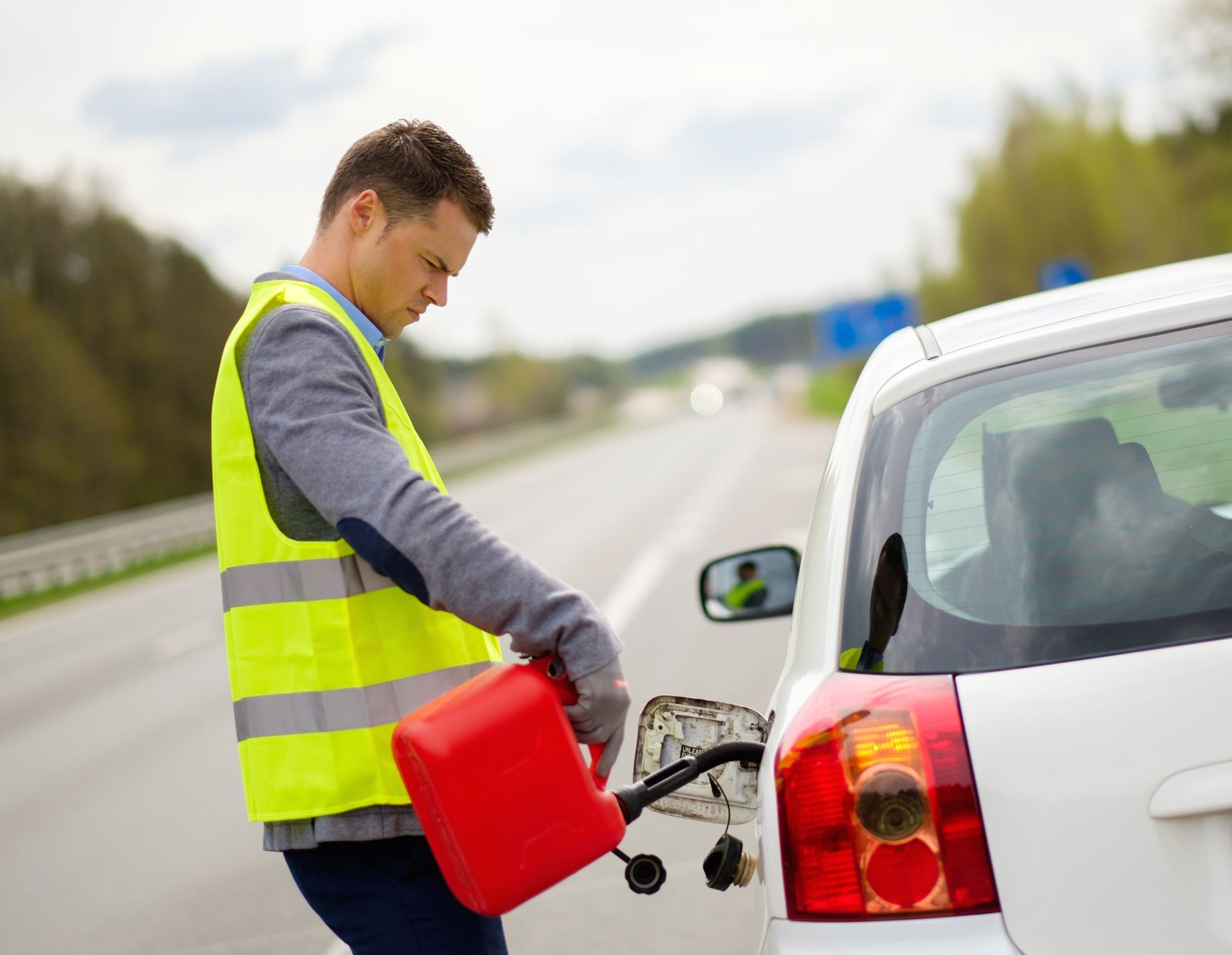 Man in a safety vest refueling a white car with a red gas can on the roadside.