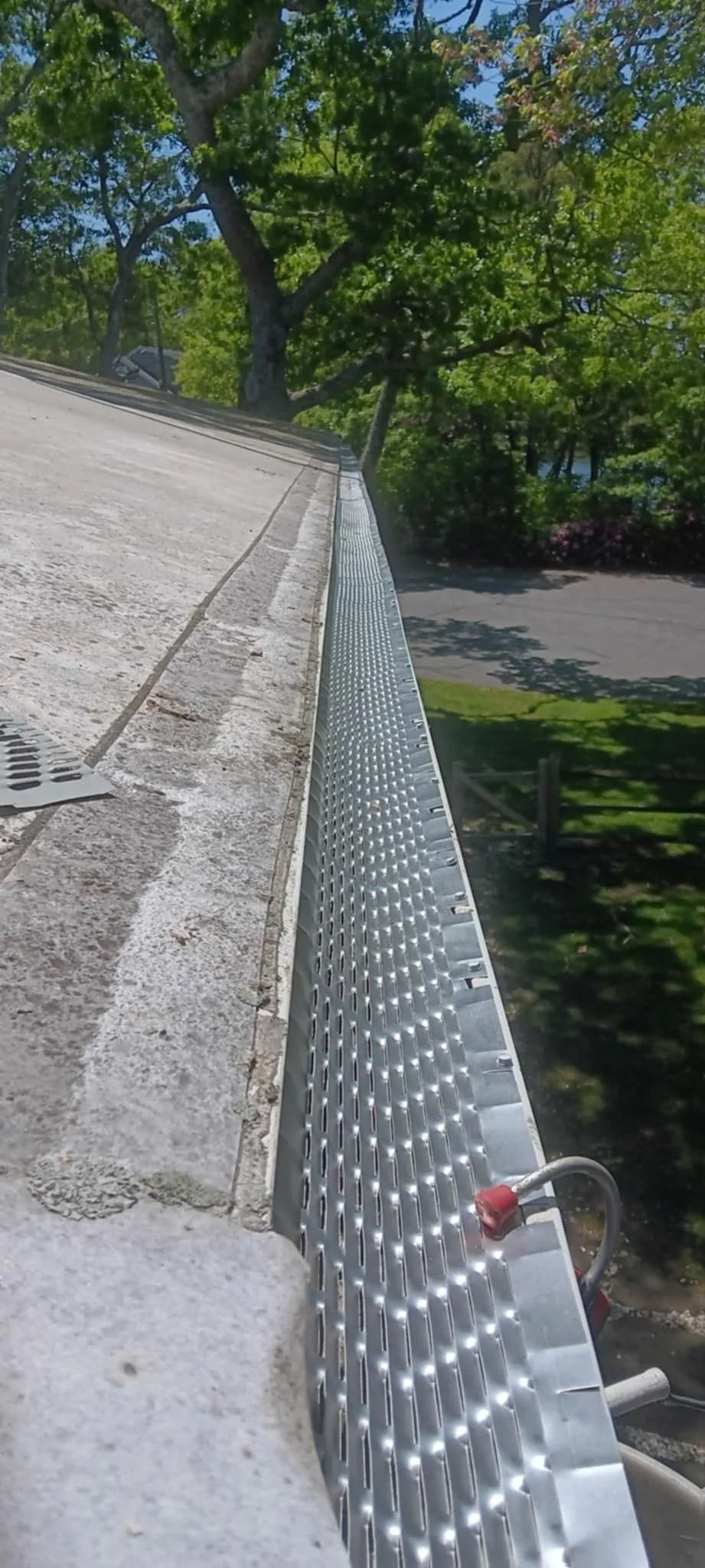 Rooftop edge with metal gutter guard beside a sloped shingle roof and leafy trees below