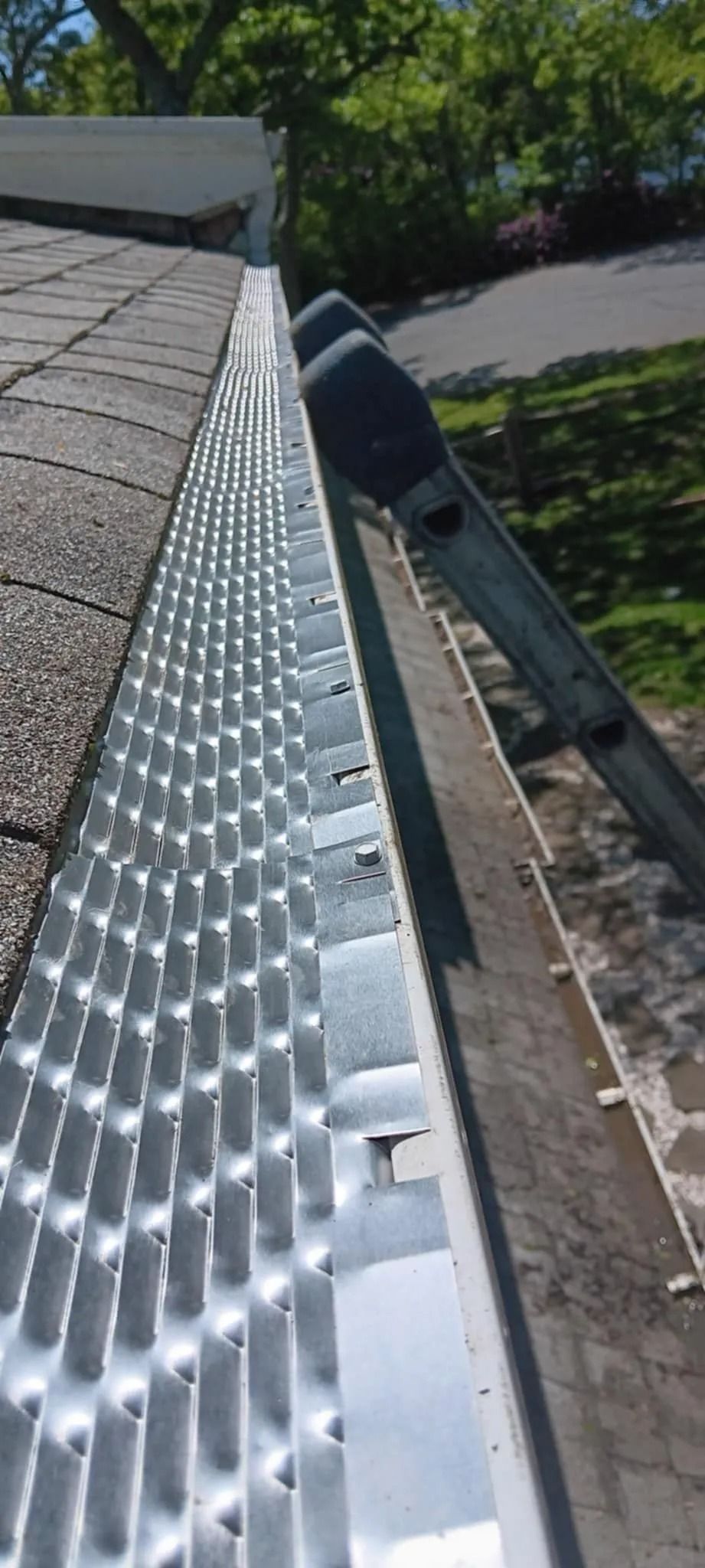 Roof gutter lined with white gutter guards beside gray shingles and trees below