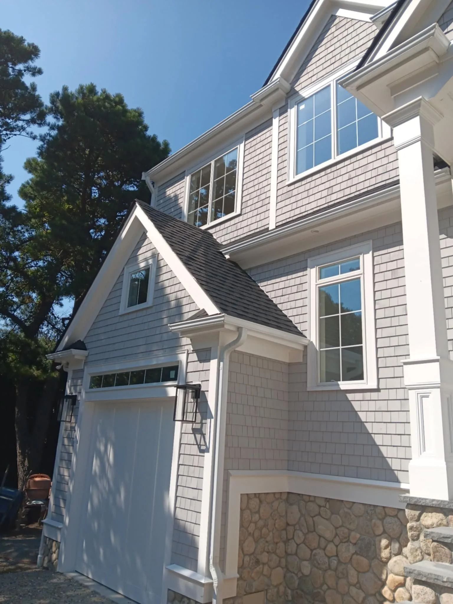 White house exterior with a garage, gabled roof, and stone foundation beside tall trees