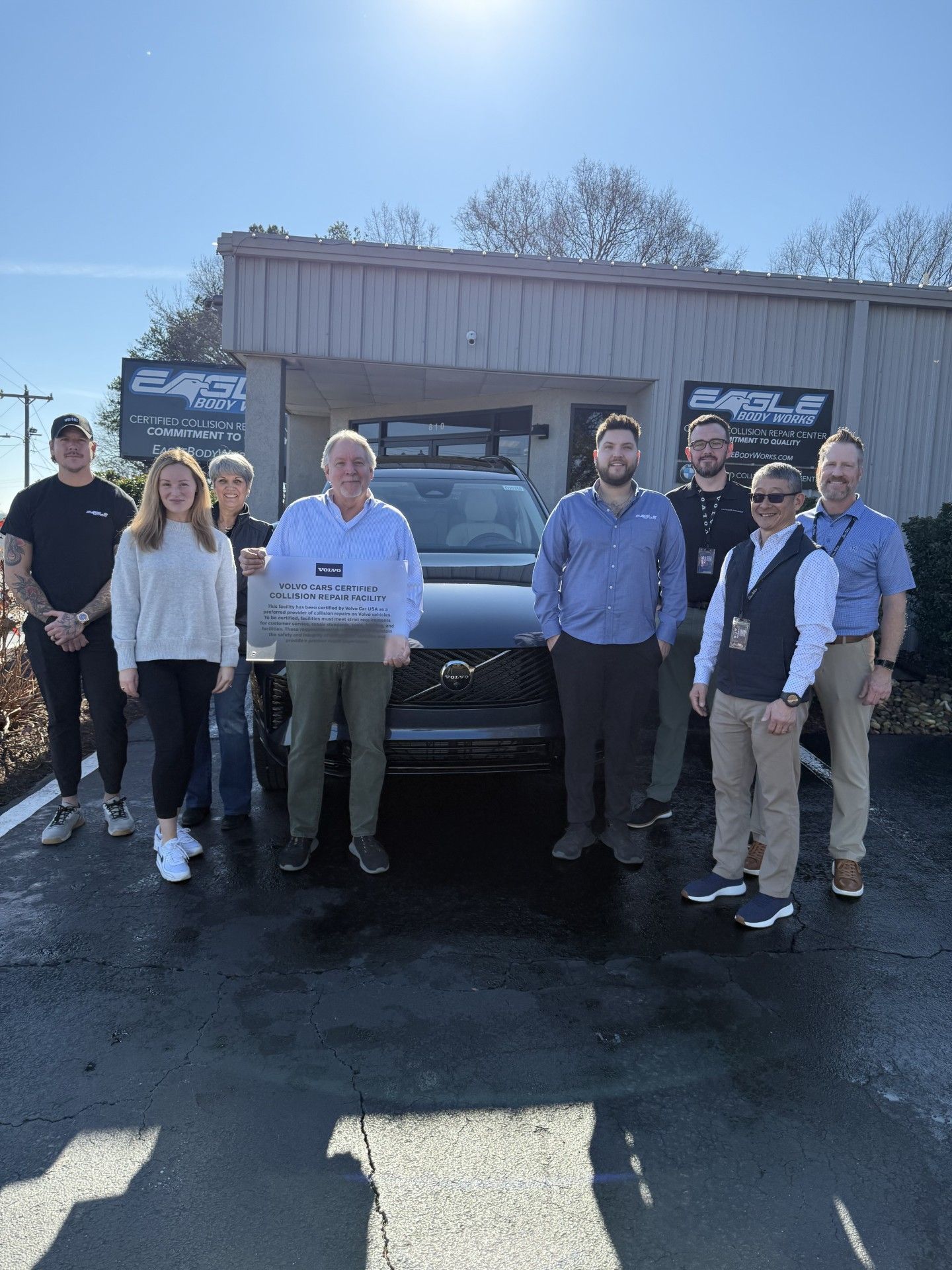 Group of people standing in front of a black SUV outside a building on a sunny day. They are holding a sign.