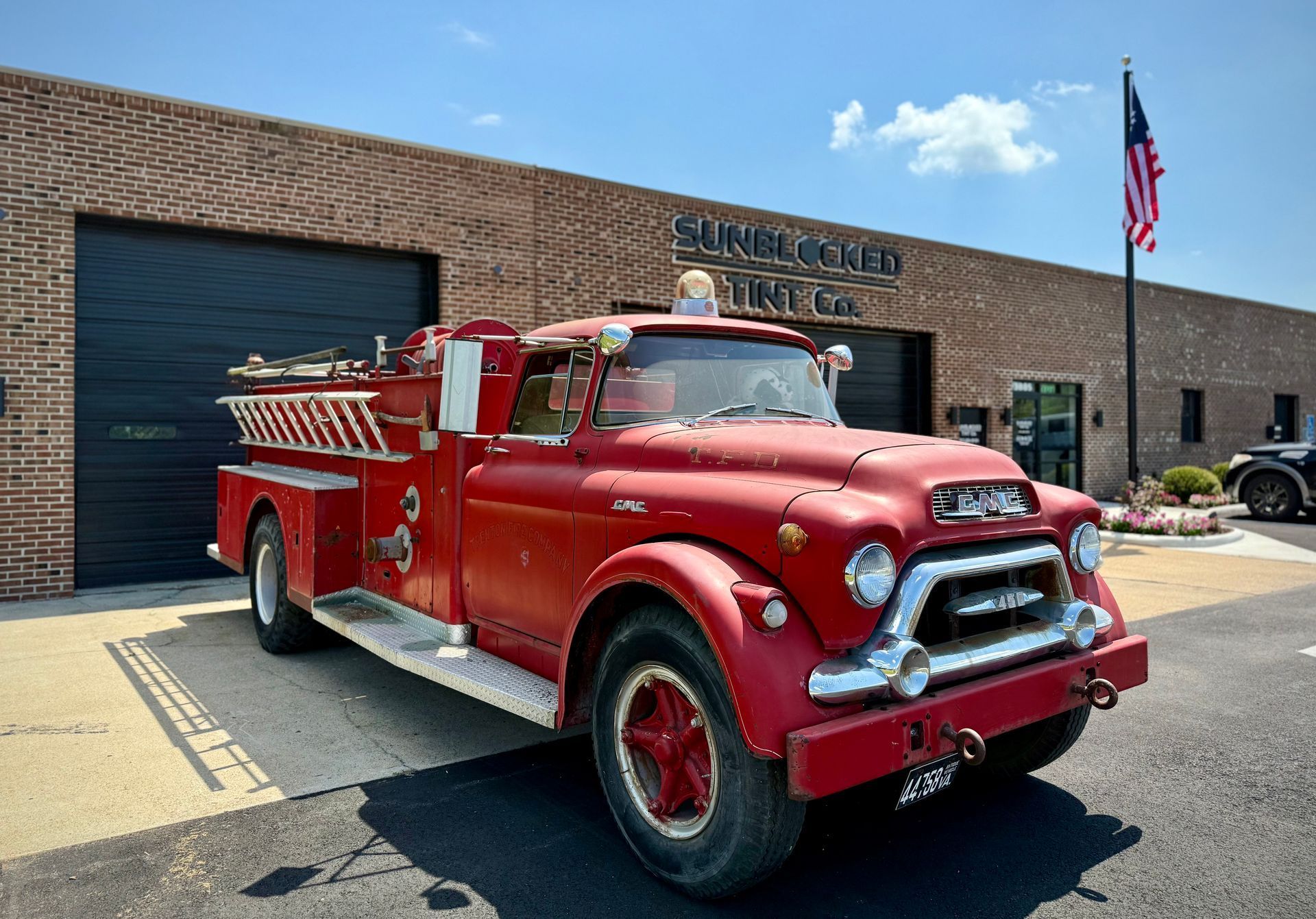 Red vintage fire truck parked in front of a brick building with a black garage door and American flag.