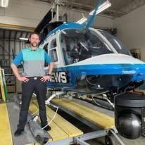 Man in front of a news helicopter. He's wearing a blue shirt. The helicopter is blue and white.
