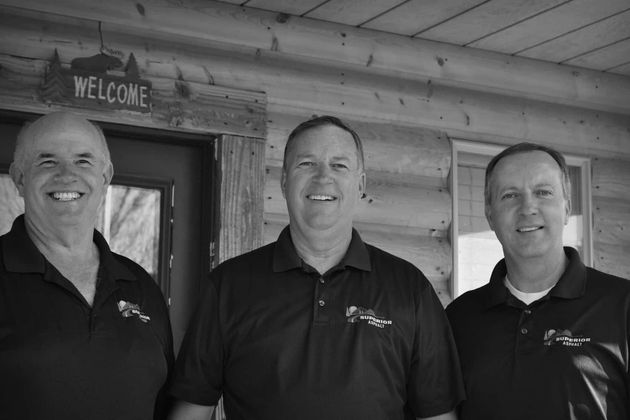 Three smiling men in black shirts stand in front of a log cabin porch.