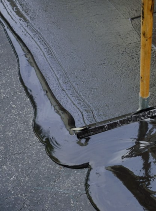 Dark liquid being spread on a gray pavement with a squeegee broom handle visible