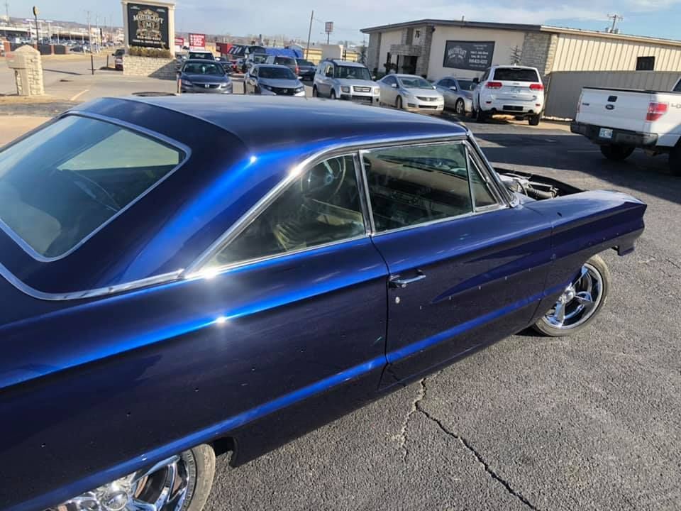 Blue classic car parked in an asphalt lot. Chrome wheels and shiny paint. Buildings in the background.
