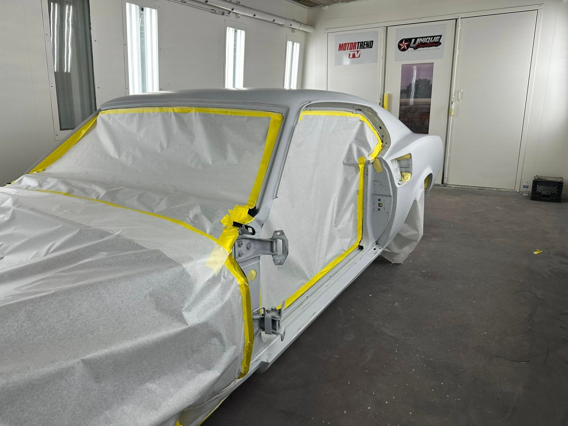 Car in a paint booth, covered in plastic and yellow tape, ready for painting.