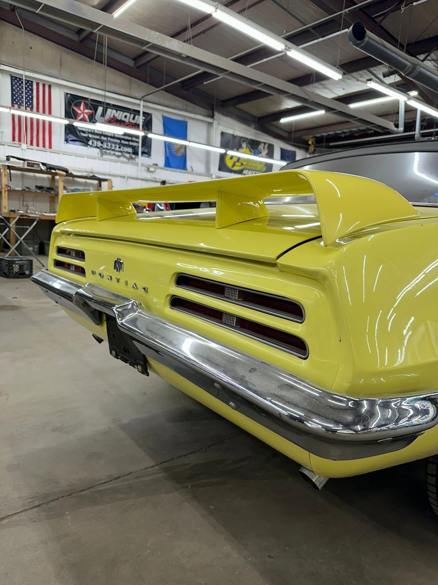 Yellow classic car's rear with spoiler and chrome bumper, indoors under shop lights.