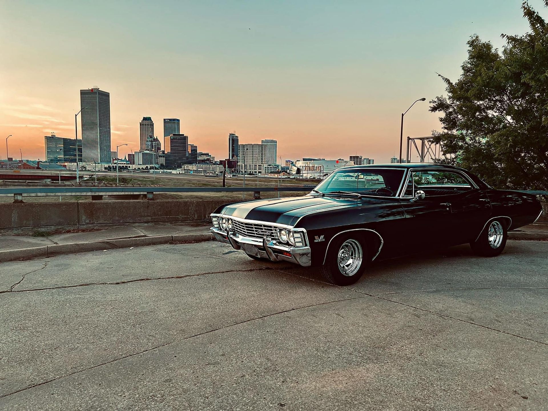 Black vintage car parked on road with cityscape and sunset in the background.