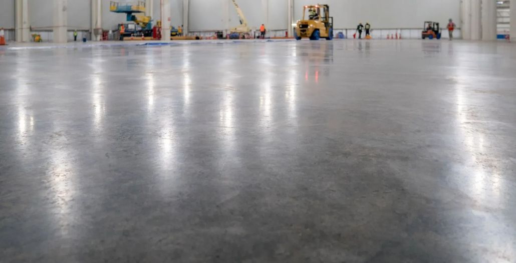 Large, polished concrete floor in a warehouse with construction equipment and workers in the background.