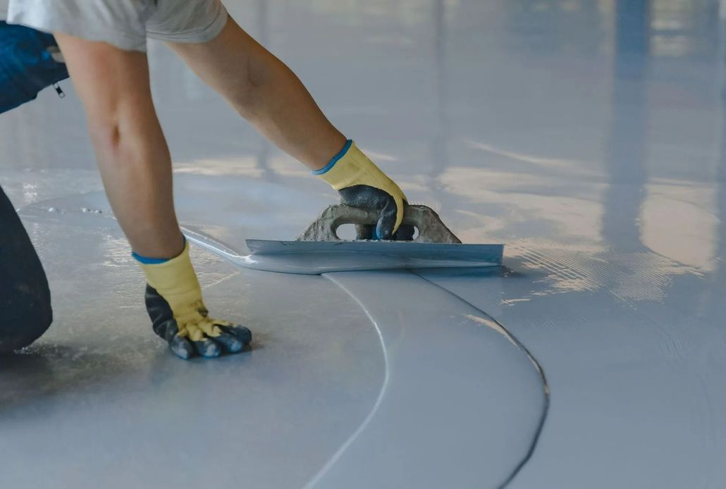 Person using a trowel to smooth epoxy flooring, wearing gloves, in a light-colored interior setting.