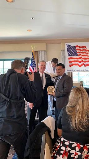 Man in suit holding award, posing with another man in suit indoors with US flag.