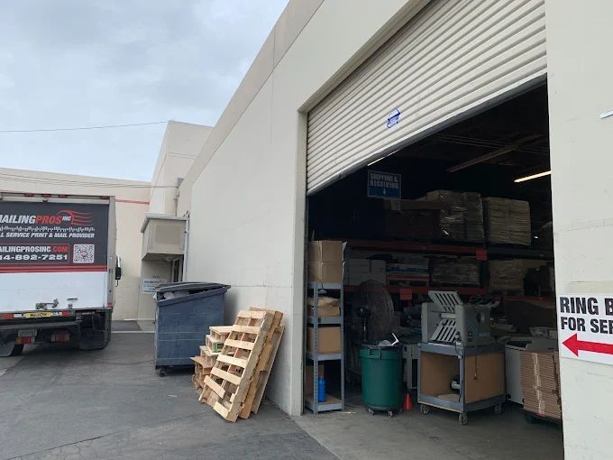 Warehouse exterior with open bay door, truck, pallets, and recycling bin.