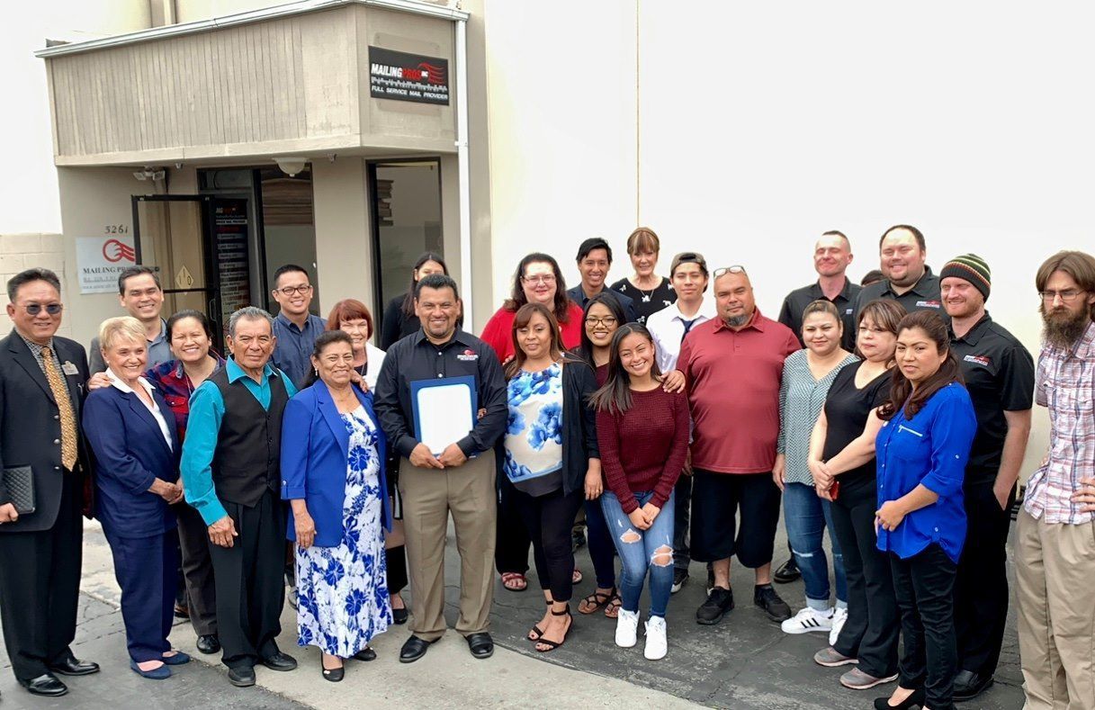 Group of people posing in front of a building. Some are smiling. The building is light-colored.