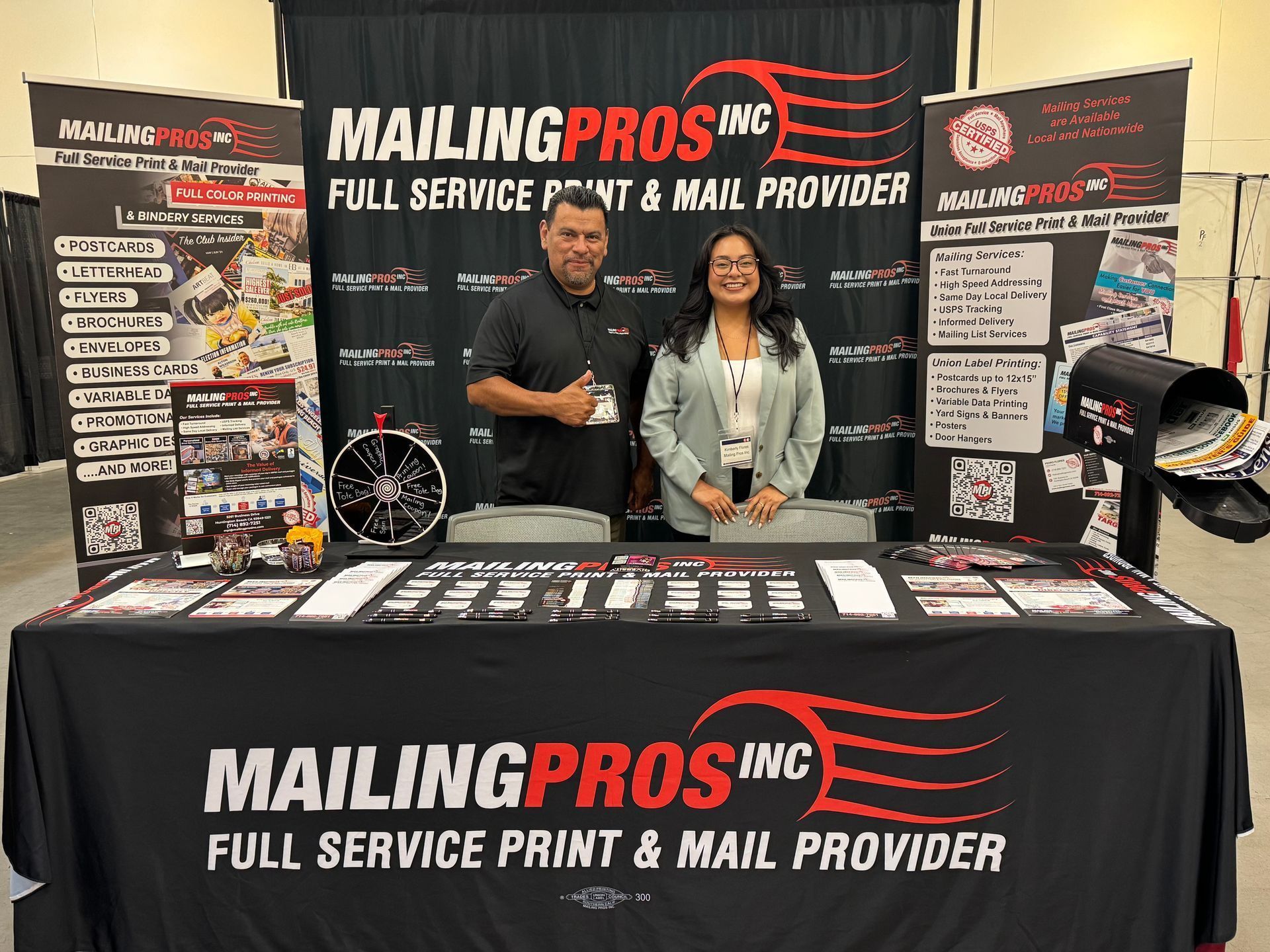 Two people stand behind a trade show booth for "Mailing Pros Inc," offering print and mail services.